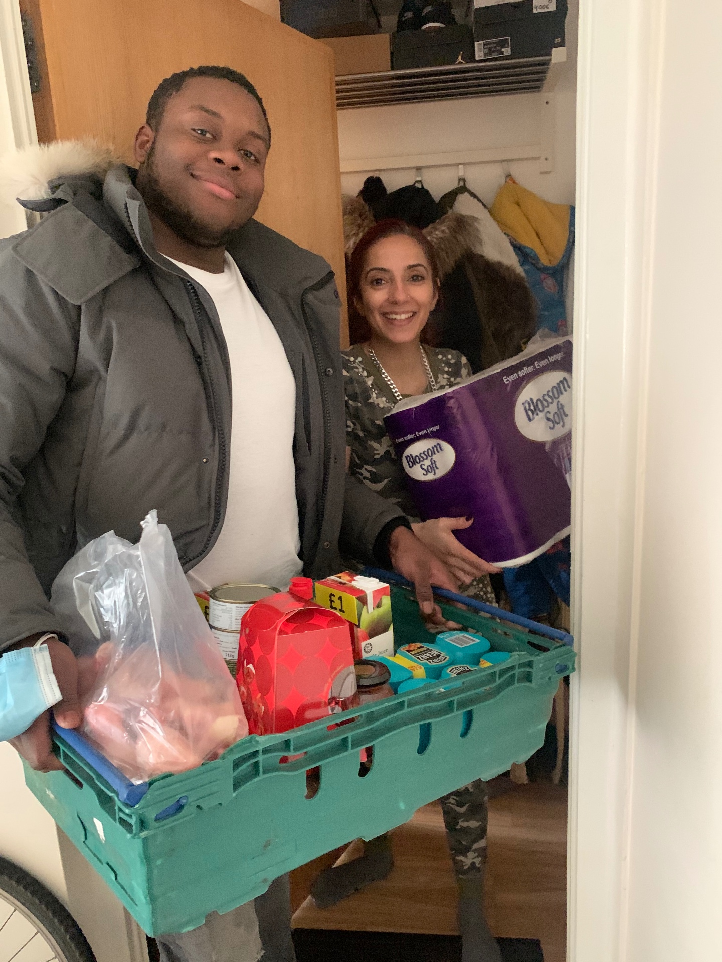 Two volunteers at a doorstep holding a green crate of fresh food and household essentials